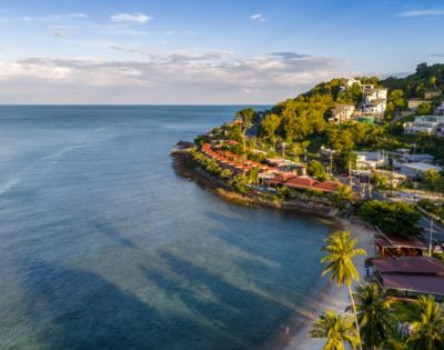 Aerial view of a coastal landscape with lush green hills, red-roofed houses, and a calm blue sea. Palm trees line a sandy beach, creating a serene, tropical vibe.