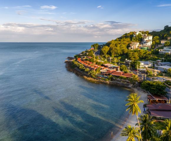 Aerial view of a coastal landscape with lush green hills, red-roofed houses, and a calm blue sea. Palm trees line a sandy beach, creating a serene, tropical vibe.