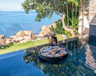 A woman in a hat sits at an infinity pool's edge, enjoying a floating breakfast with dishes. Overlooks a serene ocean view and lush greenery.