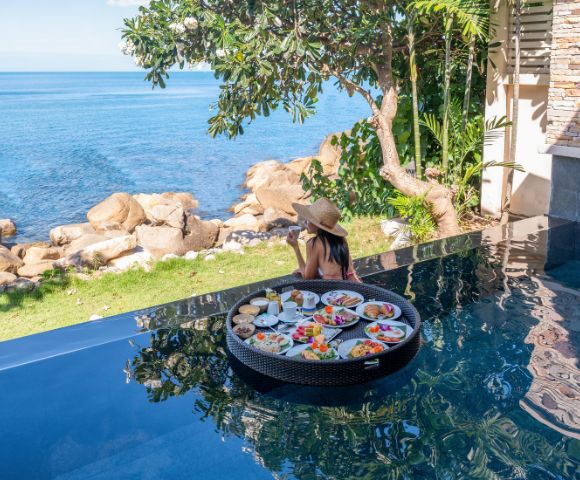A woman in a hat sits at an infinity pool's edge, enjoying a floating breakfast with dishes. Overlooks a serene ocean view and lush greenery.