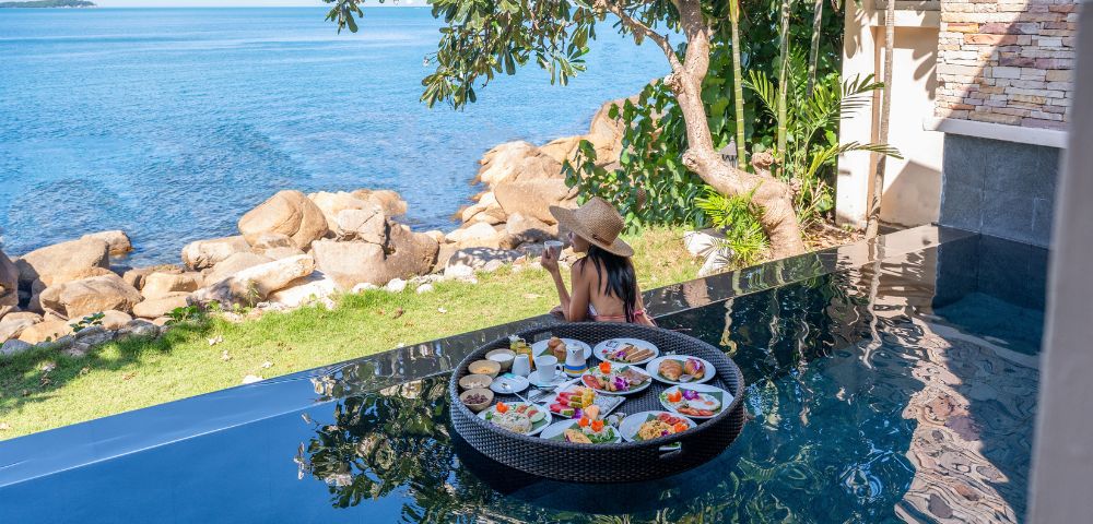 A woman in a hat sits at an infinity pool's edge, enjoying a floating breakfast with dishes. Overlooks a serene ocean view and lush greenery.