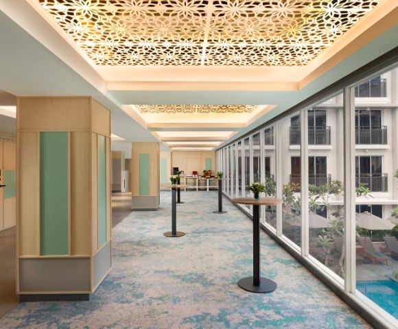 Spacious hallway with patterned ceiling and large windows overlooking a courtyard. Blue-green carpet and minimalist décor create a serene, modern ambiance.