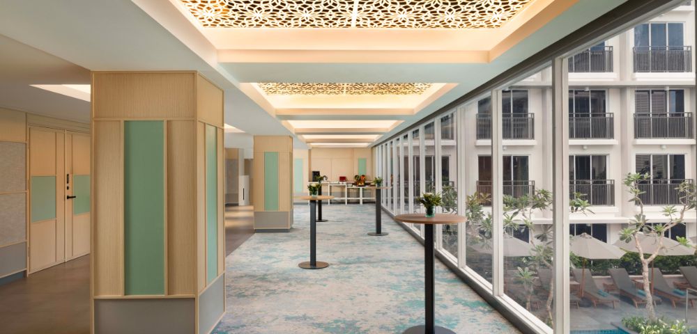 Spacious hallway with patterned ceiling and large windows overlooking a courtyard. Blue-green carpet and minimalist décor create a serene, modern ambiance.
