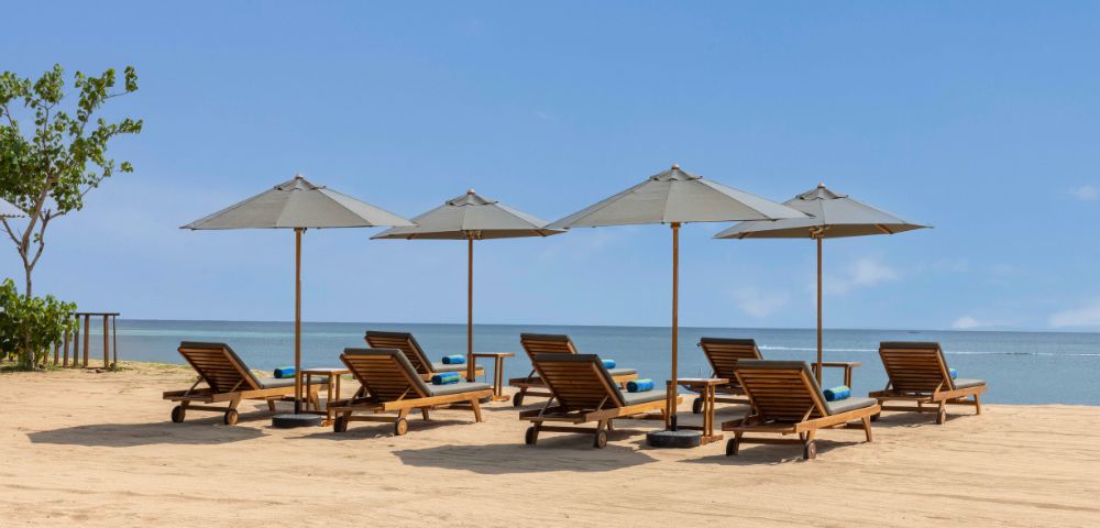 Six wooden loungers with blue cushions are arranged under gray umbrellas on a sandy beach, facing a calm ocean under a clear blue sky.