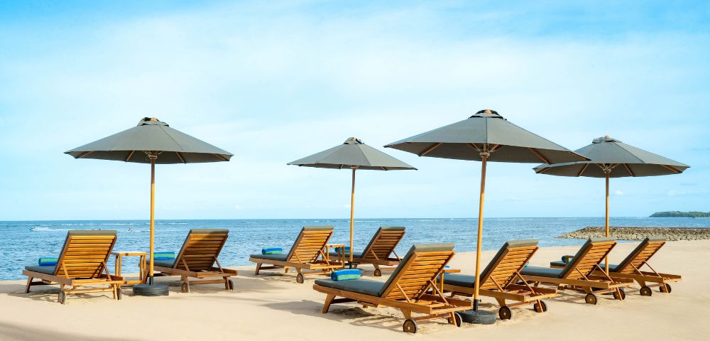Sandy beach with wooden lounge chairs and grey umbrellas facing a calm blue ocean under a clear sky; a serene and inviting atmosphere.
