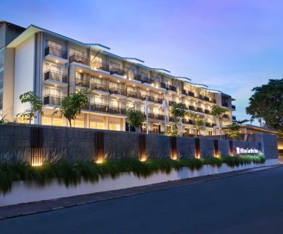 Modern hotel facade at dusk with warm lighting. Multiple balconies and well-lit exterior create a welcoming atmosphere, flanked by greenery and a calm street.