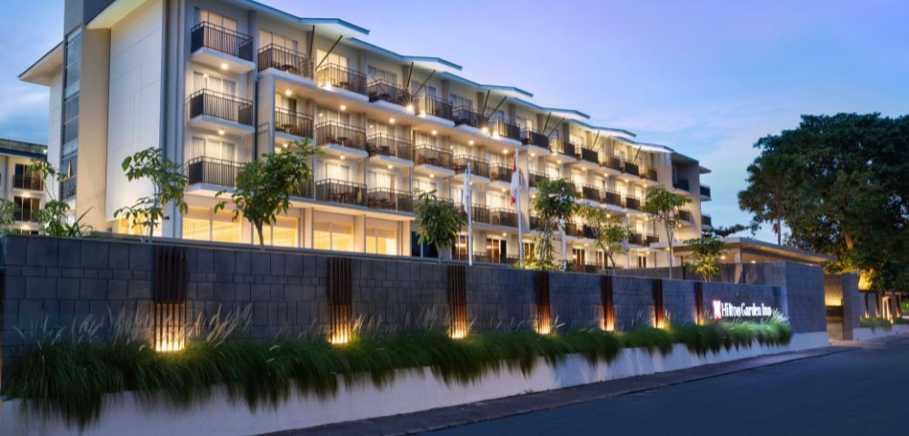 Modern hotel facade at dusk with warm lighting. Multiple balconies and well-lit exterior create a welcoming atmosphere, flanked by greenery and a calm street.
