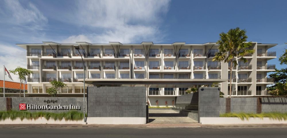Four-story Hilton Garden Inn hotel exterior with modern design, large windows, and a welcoming atmosphere under a clear blue sky.