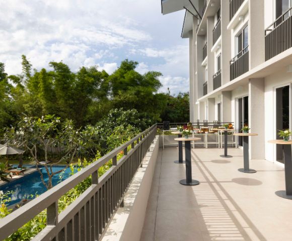 Balcony with tall tables overlooks a lush garden and pool under a partly cloudy sky. Tall trees provide a serene, refreshing atmosphere.