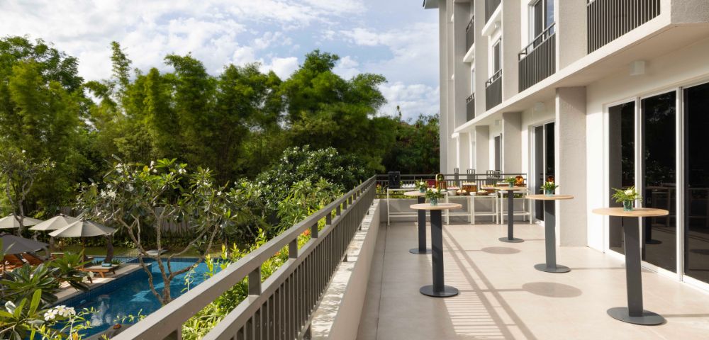 Balcony with tall tables overlooks a lush garden and pool under a partly cloudy sky. Tall trees provide a serene, refreshing atmosphere.