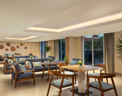 Modern lounge with wooden tables and blue cushioned chairs, adorned with potted plants. Large windows offer natural light, creating a cozy, inviting atmosphere.