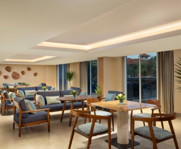 Modern lounge with wooden tables and blue cushioned chairs, adorned with potted plants. Large windows offer natural light, creating a cozy, inviting atmosphere.