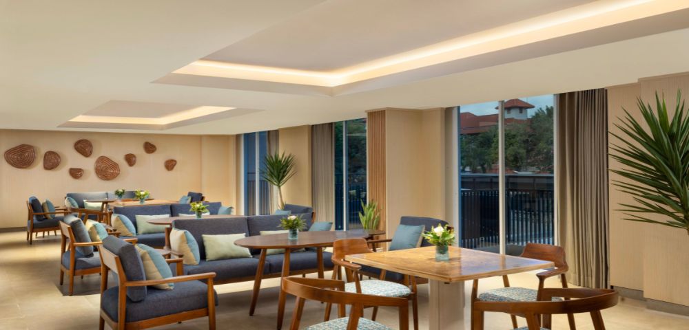 Modern lounge with wooden tables and blue cushioned chairs, adorned with potted plants. Large windows offer natural light, creating a cozy, inviting atmosphere.