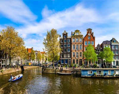 Scenic view of Amsterdam canal with charming, narrow buildings and vibrant trees on a sunny day. A small boat glides through the calm water.
