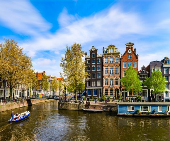 Scenic view of Amsterdam canal with charming, narrow buildings and vibrant trees on a sunny day. A small boat glides through the calm water.