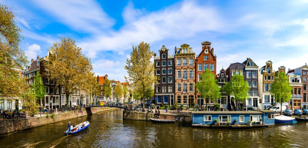 Scenic view of Amsterdam canal with charming, narrow buildings and vibrant trees on a sunny day. A small boat glides through the calm water.