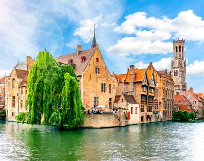 Charming canal in Bruges, bordered by historic brick buildings and lush greenery. A tall clock tower rises in the background under a bright blue sky.