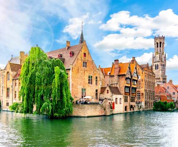 Charming canal in Bruges, bordered by historic brick buildings and lush greenery. A tall clock tower rises in the background under a bright blue sky.