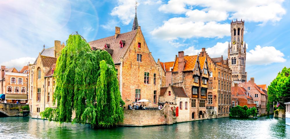 Charming canal in Bruges, bordered by historic brick buildings and lush greenery. A tall clock tower rises in the background under a bright blue sky.