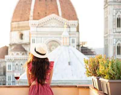 A woman in a pink dress and sunhat gazes at the Florence Cathedral from a terrace at sunset, a glass of red wine beside her, creating a serene ambiance.