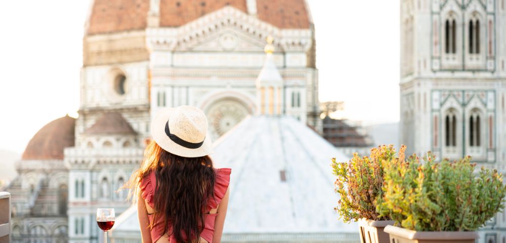 A woman in a pink dress and sunhat gazes at the Florence Cathedral from a terrace at sunset, a glass of red wine beside her, creating a serene ambiance.