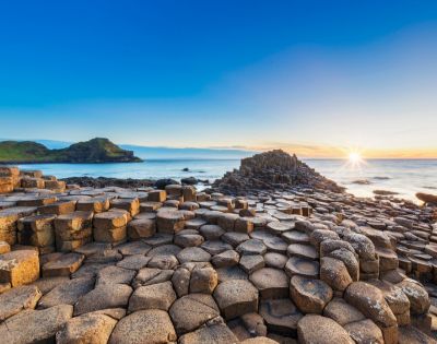 Sunlit scene of Giant's Causeway with hexagonal basalt columns leading to the ocean under a clear blue sky at sunset, evoking awe and tranquility.
