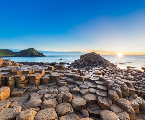 Sunlit scene of Giant's Causeway with hexagonal basalt columns leading to the ocean under a clear blue sky at sunset, evoking awe and tranquility.
