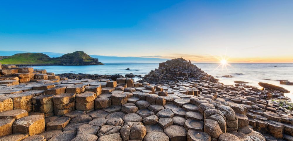 Sunlit scene of Giant's Causeway with hexagonal basalt columns leading to the ocean under a clear blue sky at sunset, evoking awe and tranquility.