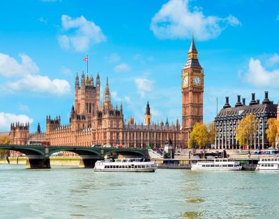 Iconic view of London's Big Ben and the Houses of Parliament under a blue sky. The Thames River flows with white boats, evoking a lively, historic ambiance.