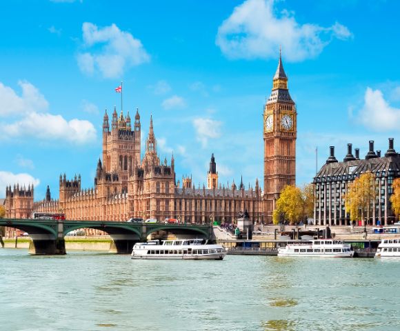 Iconic view of London's Big Ben and the Houses of Parliament under a blue sky. The Thames River flows with white boats, evoking a lively, historic ambiance.