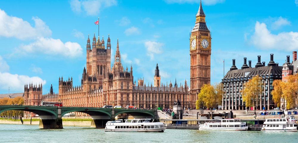 Iconic view of London's Big Ben and the Houses of Parliament under a blue sky. The Thames River flows with white boats, evoking a lively, historic ambiance.