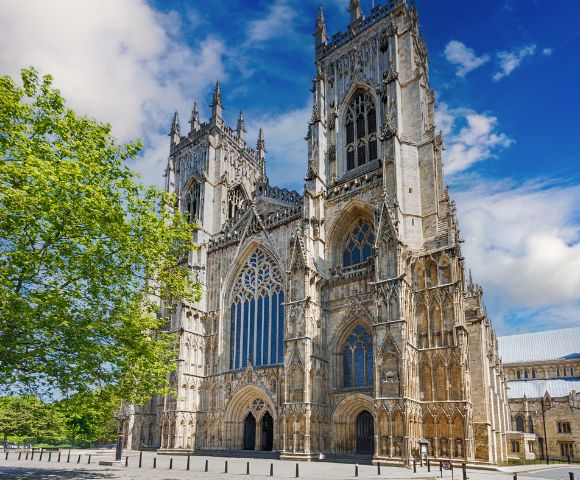 Gothic cathedral under a vibrant blue sky, adorned with intricate spires and arched windows. A lush green tree adds contrast, conveying serenity and grandeur.