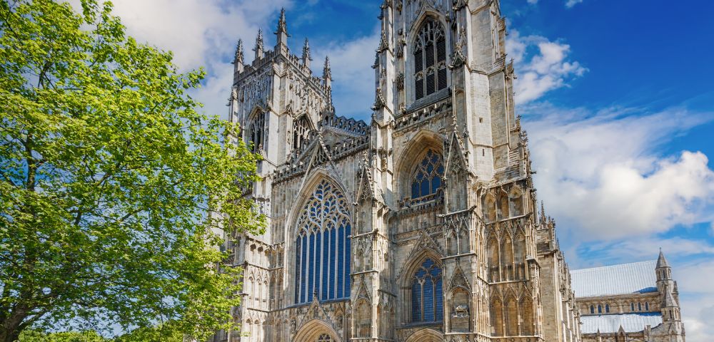 Gothic cathedral under a vibrant blue sky, adorned with intricate spires and arched windows. A lush green tree adds contrast, conveying serenity and grandeur.