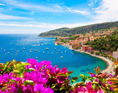 A vibrant coastal scene with bright magenta flowers in the foreground, overlooking a curving bay dotted with boats and surrounded by lush hills and colorful buildings.