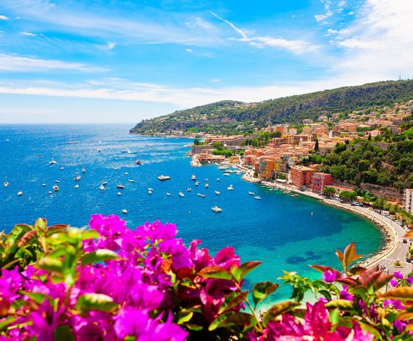 A vibrant coastal scene with bright magenta flowers in the foreground, overlooking a curving bay dotted with boats and surrounded by lush hills and colorful buildings.