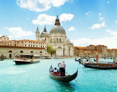 Gondolas glide on vibrant blue waters in Venice under a sunny sky. The grand, domed Basilica di Santa Maria della Salute stands prominently in the background.