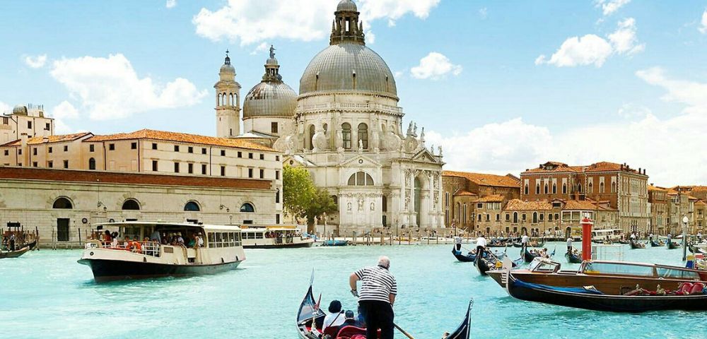 Gondolas glide on vibrant blue waters in Venice under a sunny sky. The grand, domed Basilica di Santa Maria della Salute stands prominently in the background.