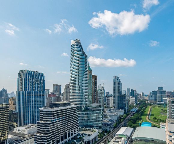 A vibrant cityscape with modern skyscrapers under a blue, partly cloudy sky. A mix of tall buildings frames a green park, conveying a dynamic urban atmosphere.
