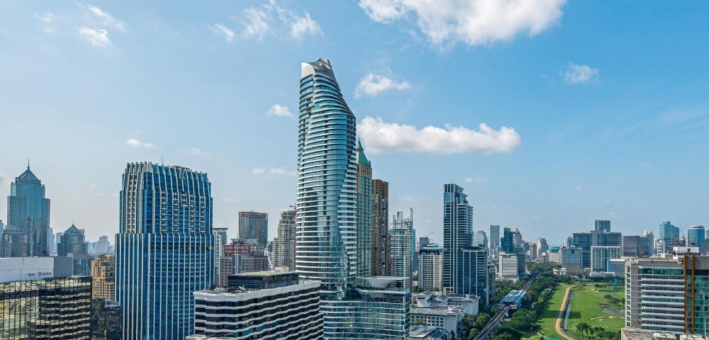 A vibrant cityscape with modern skyscrapers under a blue, partly cloudy sky. A mix of tall buildings frames a green park, conveying a dynamic urban atmosphere.