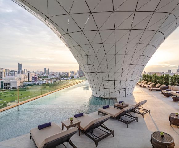 Rooftop infinity pool with sleek lounge chairs beneath a modern, geometric canopy. Cityscape view in the distance, soft sunrise lighting.