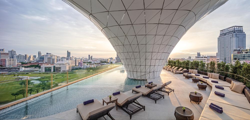 Rooftop infinity pool with sleek lounge chairs beneath a modern, geometric canopy. Cityscape view in the distance, soft sunrise lighting.