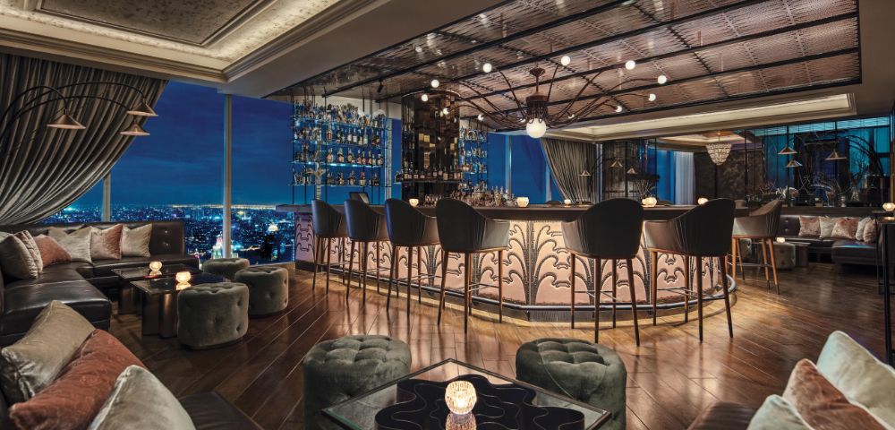 Luxurious rooftop bar with elegant decor, featuring plush seating and a curved bar, overlooking a cityscape at night. Warm, inviting ambiance.
