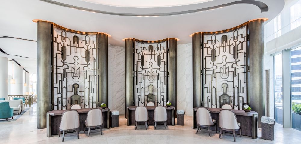 Elegant hotel lobby with modern decor features three artistic, geometric wall panels. Curved desks and chairs add sophistication, while lighting sets a calming tone.