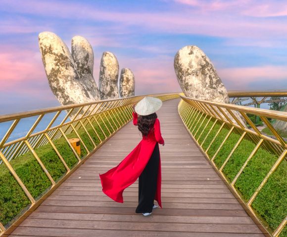 A person in a long red dress and traditional conical hat walks along a golden pedestrian bridge framed by giant stone hand sculptures. The sky is pink and blue at sunset, casting a warm glow over the scenic landscape.