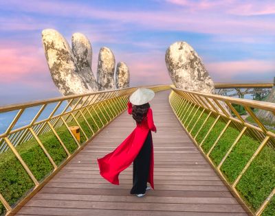 A person in a long red dress and traditional conical hat walks along a golden pedestrian bridge framed by giant stone hand sculptures. The sky is pink and blue at sunset, casting a warm glow over the scenic landscape.