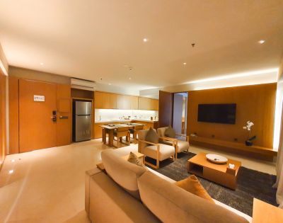 Spacious, modern living room with warm lighting. Features a sofa, armchairs, TV, coffee table, and a kitchen area with wooden cabinetry and stainless steel fridge.