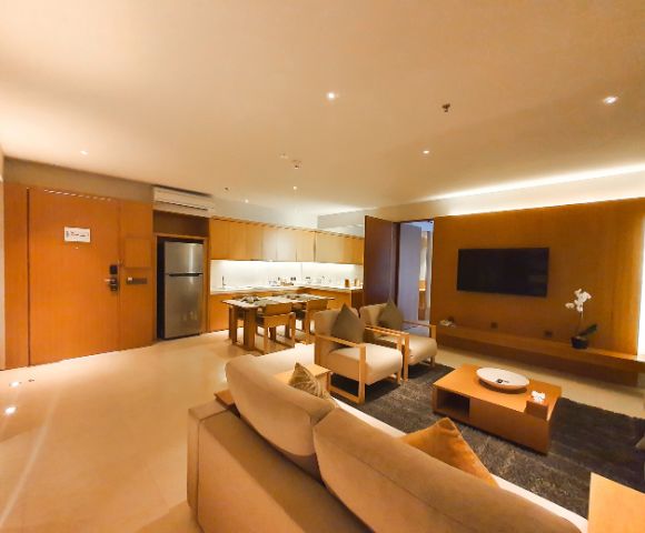 Spacious, modern living room with warm lighting. Features a sofa, armchairs, TV, coffee table, and a kitchen area with wooden cabinetry and stainless steel fridge.