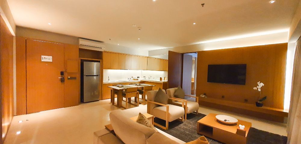 Spacious, modern living room with warm lighting. Features a sofa, armchairs, TV, coffee table, and a kitchen area with wooden cabinetry and stainless steel fridge.