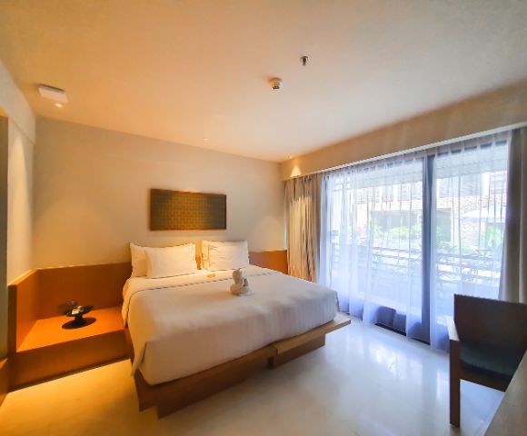 A minimalist hotel room with a king-sized bed adorned with white linens and a towel swan. Soft lighting, large window with sheer curtains, and a serene ambiance.