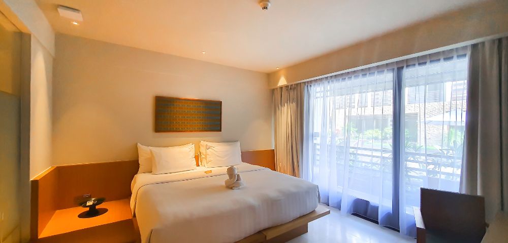 A minimalist hotel room with a king-sized bed adorned with white linens and a towel swan. Soft lighting, large window with sheer curtains, and a serene ambiance.